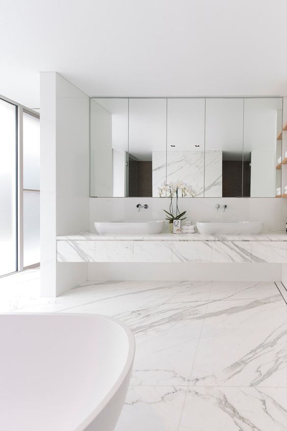a refined white minimalist bathroom with white marble tiles on the floor and a white marble vanity, white appliances and a mirror wall