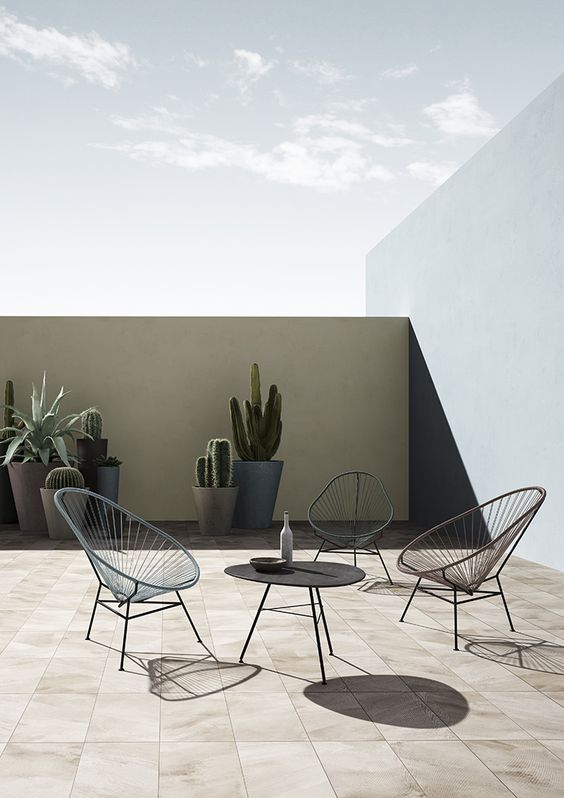 a simple and minimal terrace with a tan wall, several chairs, a sculptural table and tall planters with succulents and cacti