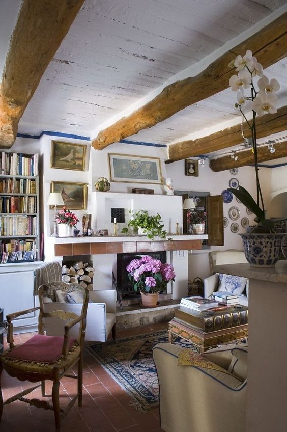 a small Provence living room with a fireplace, wooden beams, neutral furniture and stacked books plus decorative plates