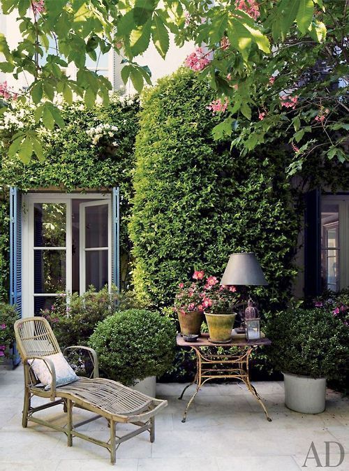 a small and delicate Provence terrace with a wooden lounger, a side table with blooms and a table lamp and lots of greenery