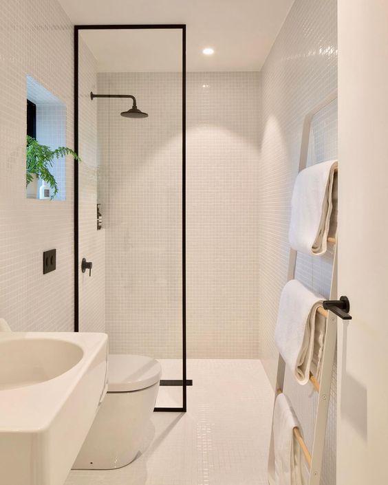 a small white minimalist bathroom clad with tiny tiles, with a ladder for towels, white appliances and a shower with a glass partition