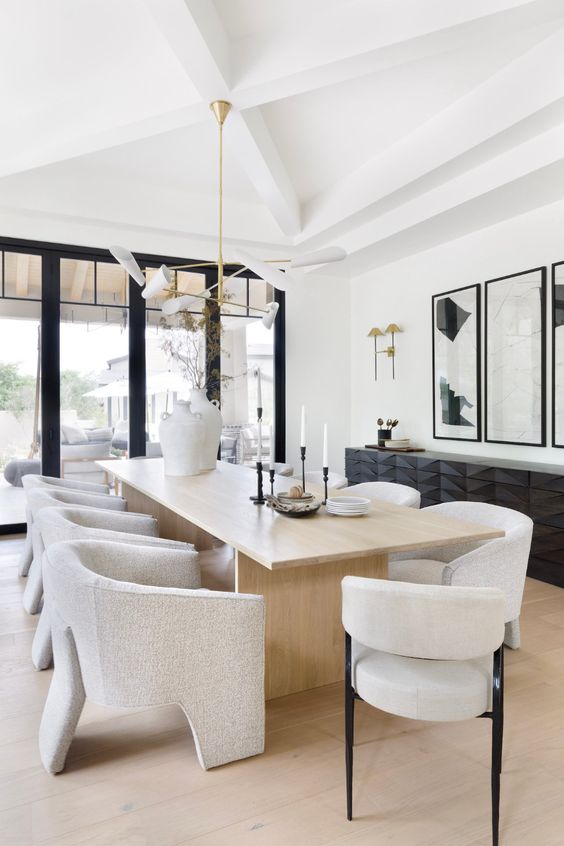A stylish and refined minimalist dining space with a black sculptural console, a light stained table and neutral chairs, a catchy chandelier
