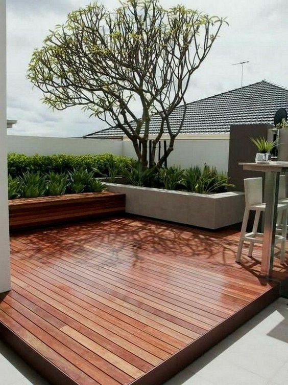 a stylish minimalist rich-stained deck with a built-in bench, potted greenery and a tree, a metal and wood tall table and tall stools