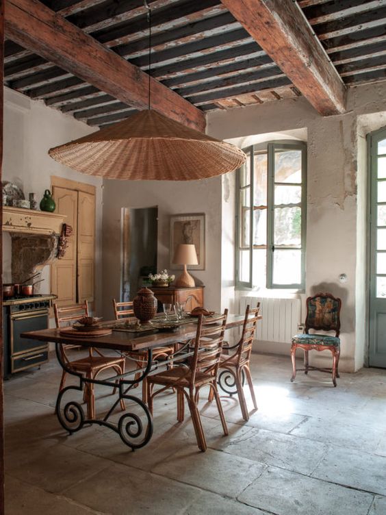 A vintage kitchen and dining space in one, with rich stained wooden beams, a refined dining table and stained chairs, a wicker pendant lamp and some furniture