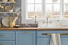 a blue farmhouse kitchen with shaker cabinets, burtcherblock countertops, open shelves and brass touches plus a window that opens on the garden