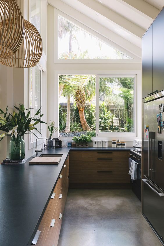 A chic modern kitchen with light stained cabinets, black stone countertops, a large attic window with a tropical garden view