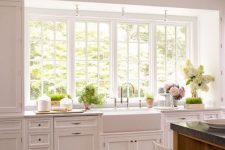 a chic white kitchen with shaker style cabinets, stone countertops, vintage fixtures and a large window that opens on the garden