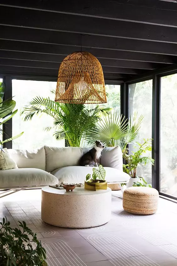 a contemporaru boho screened porch with a white sofa, a round coffee table, a jute pouf, potted greenery and a woven pendant lamp