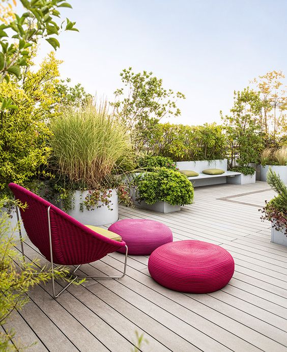 A contemporary terrace with lots of greenery, built in benches, a fuchsia wicker chair, fuchsia poufs and a green cushion