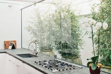 a contemporary white kitchen with a dark countertop, a glazed wall with a view of the private garden and greenery