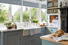 a country kitchen with serenity blue shaker style cabinets, white stone and butcherblock countertops, several windows for a garden view