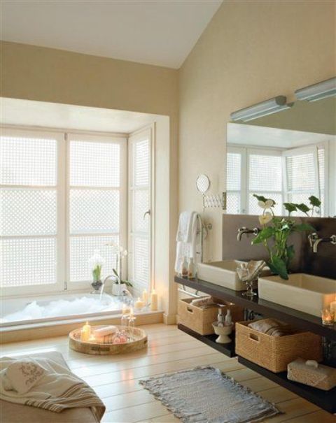 a cozy and neutral bathroom with a sunken bathtub, a wooden floor, blooms and candles in trays plus a large mirror