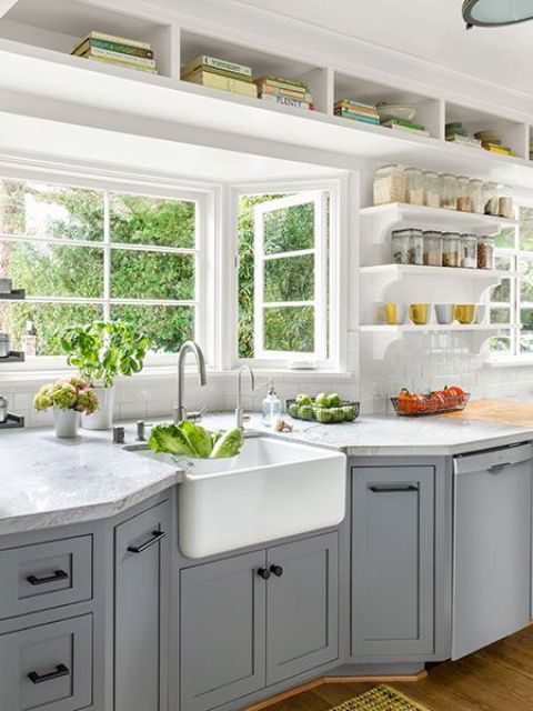 A cozy grey farmhouse kitchen with shaker style cabinets, white stone countertops, built in shelves and a bay window that opens on the garden