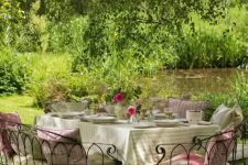 a delightful Provence outdoor space with a dining set of metal, with lots of cushions and pillows, bright blooms and greenery over it