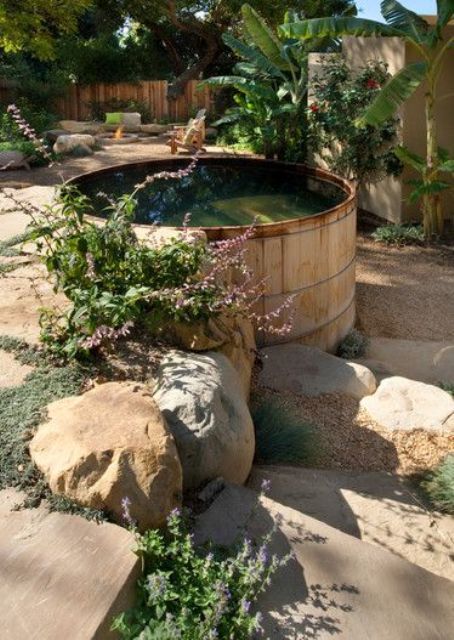 a large wooden tank is used as a plunge pool and looks very natural in rocks, greenery and blooms growing around