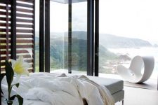 a lovely contemporary bedroom with a bed, a bench, nightstands, glass walls and a balcony plus amazing ocean views