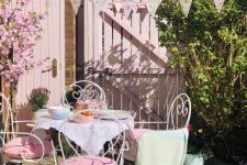 a lovely feminine terrace wiht white forged furniture, blush painted doors and pink pillows, a floral bunting and potted blooms