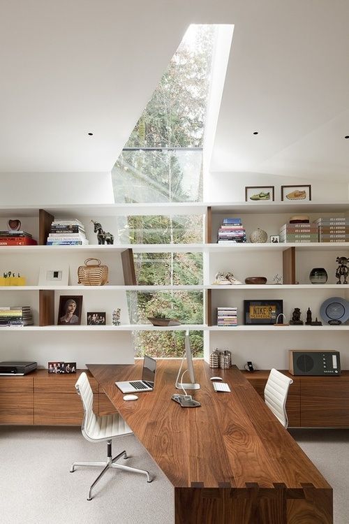 A modern home office with a catchy irregular window to enjoy garden views, lots of wall mounted shelves, sleek storage units and a built in desk