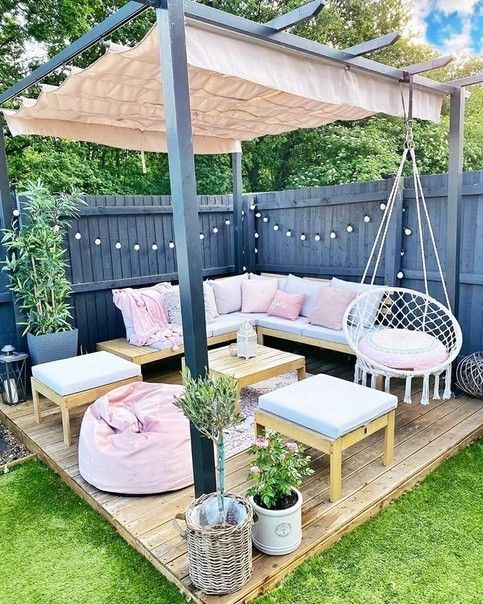 a pretty and lovely terrace wiht a wooden deck, a corner sofa with pastel pillows and blankets, a pink pouf, side tables and potted blooms