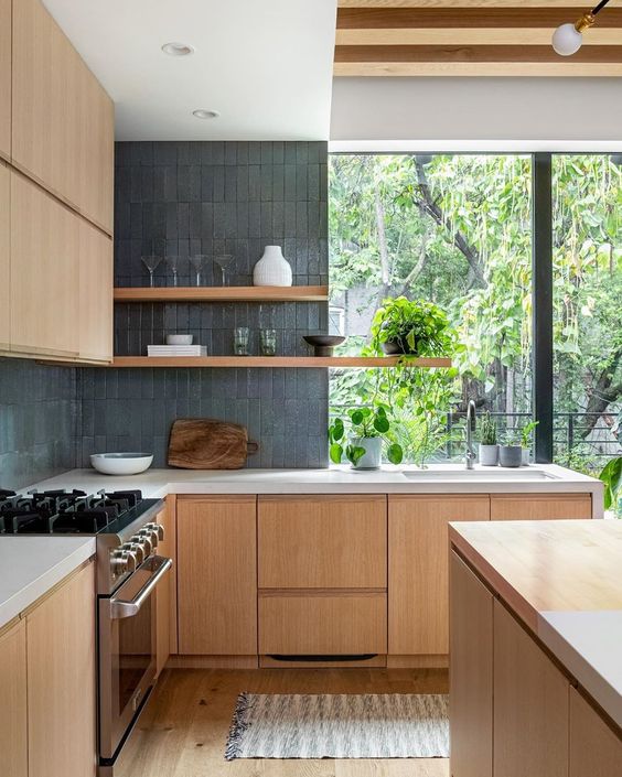 A pretty contemporary kitchen with sleek light stained cabinetry, grey skinny tiles, open shelves and a large window with greeneyr views