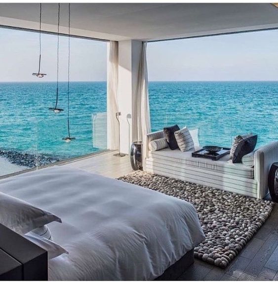 a sea bedroom with glass walls and amazing views, chic white furniture, pendant lamps and a pebble rug is fantastic