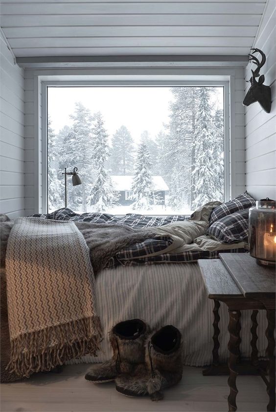 a small chalet bedroom with a glass wall, a bed with plaid bedding, a vintage wooden nightstand and a view of the forest is veyr cozy