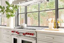 a white vintage farmhouse kitchen with shaker style cabinets, a large metal hood and a big window that opens on the garden
