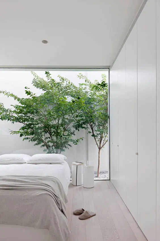an airy minimalist bedroom in neutrals, with a large bed, white nightstands and a glass wall with a view of the private courtyard