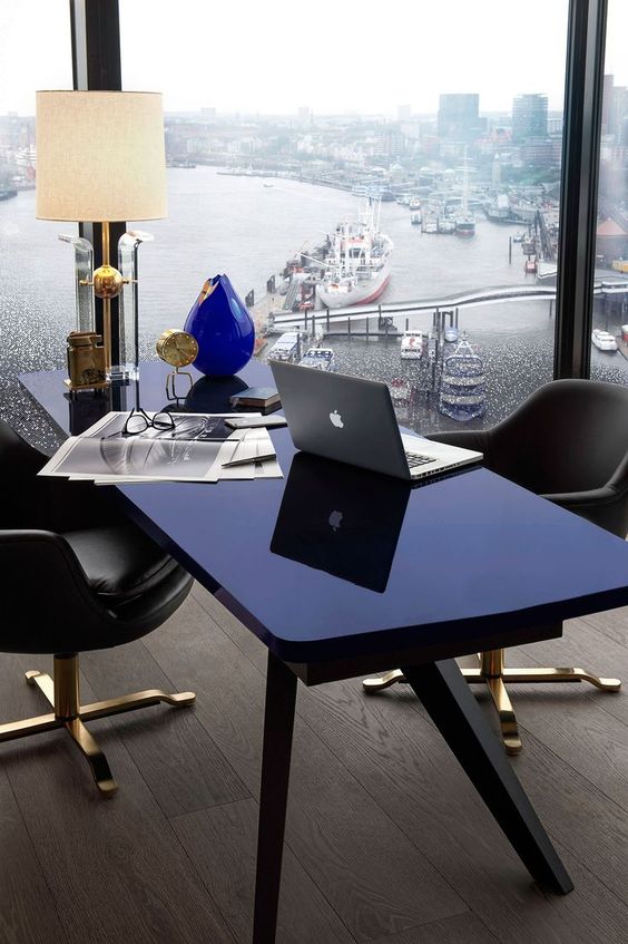 an ultra-modern home office with a glazed wall and a view of the harbor, a navy desk, grey chairs, a chic table lamp and vases