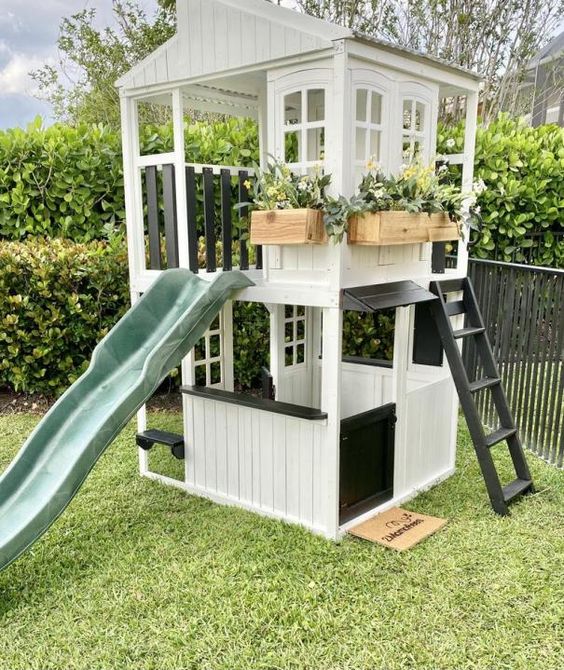 a black and white kids' playhouse with a ladder and a green slide, with potted greenery and blooms will be loved by your kids