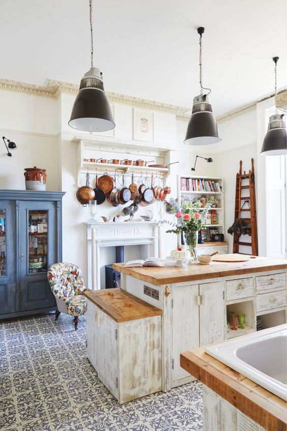 a bright shabby chic kitchen with white shabby furniture, a grey armoire, a hearth and lots of pans hanging over it