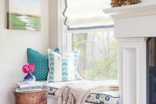 a cool and chic window nook with a Roman shade, a windowsill daybed with printed bedding and a small side table and blooms