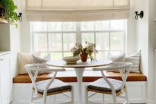 a cozy farmhouse dining space with a creamy Roman shade, a windowsill seat, a round table and woven chairs is lovely