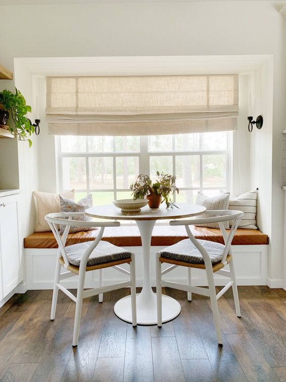 a cozy farmhouse dining space with a creamy Roman shade, a windowsill seat, a round table and woven chairs is lovely