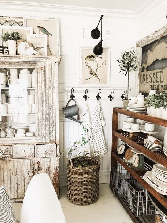 a farmhouse shabby chic kitchen with a stained open storage unit, white shabby furniture, greenery in pots and vintage artworks
