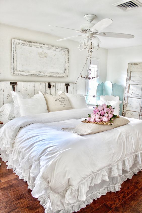 a gorgeous shabby chic bedroom in white, with a wooden bed and an artwork, a mint screen, a fan, neutral bedding with ruffles