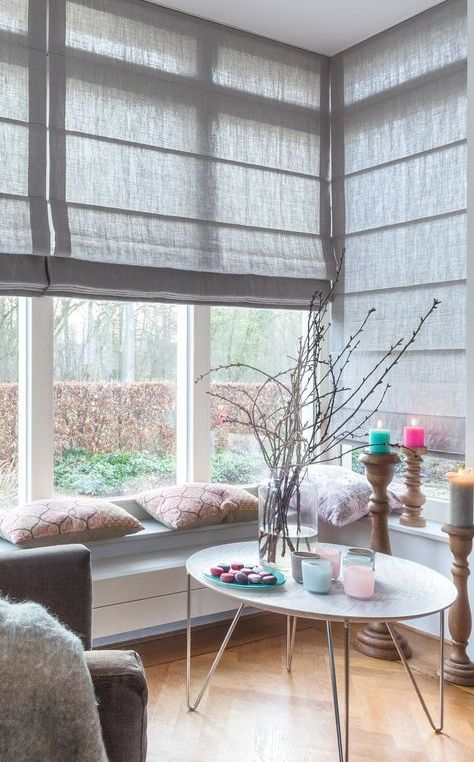 a lovely and cozy nook with a corner window, grey Roman shades, windowsill benches, a round table and bright touches