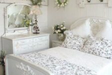 a lovely white shabby chic bedroom with refined furniture, floral bedding, a mirror on the wall, white blooms and a floral lamp