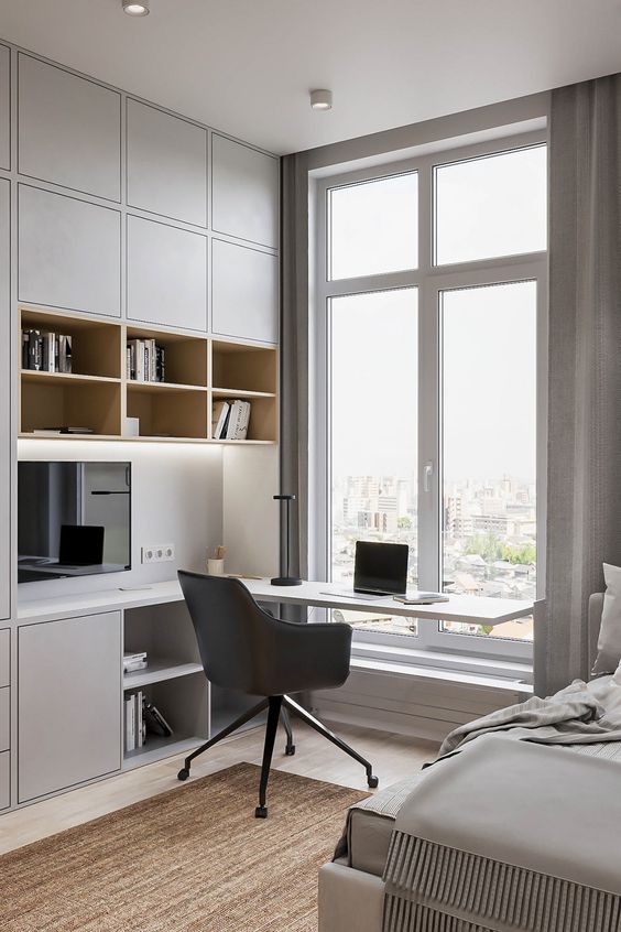 A minimalist home office nook with sleek grey storage units and built in lights, a built in desk and a black chair plus a view of the city