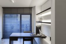 a minimalist home office with an open shelving unit with lights, a built-in desk and a blakc chair plus a floor to ceiling window