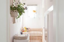 a narrow nursery in white, with a stained crib, a white dresser, some windows and artwork, greenery and toys