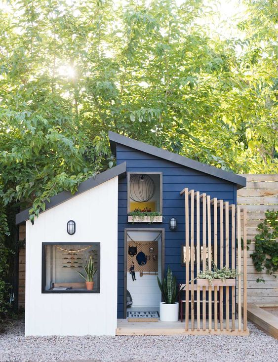 A navy playhouse with a planked porch and a wall mounted seat, potted greenery looks like a real mid century modern house