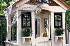a neutral kids’ playhouse with a roof, with black window frames, potted plants and blooms styled for Halloween