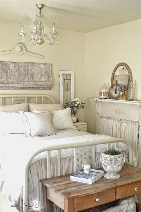 a neutral shabby chic bedroom with buttermilk walls, a metal bed, wooden furniture and a non-working fireplace and a crystal chandelier