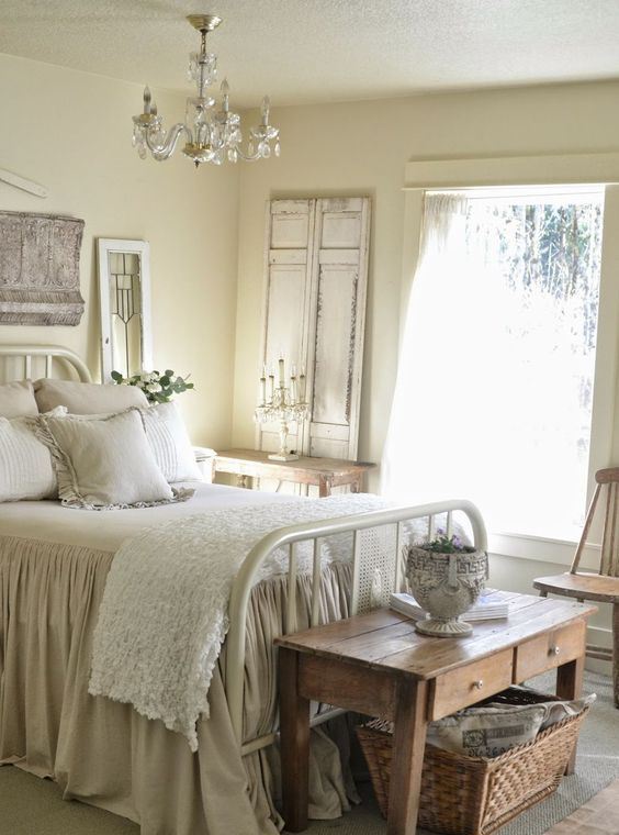 a neutral shabby chic bedroom with buttermilk walls, a white forged bed, wooden furniture, a crystal chandelier and ruffle bedding