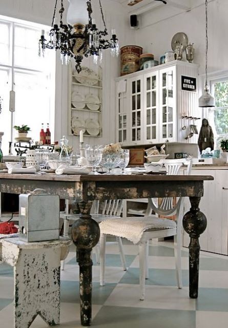 a neutral shabby chic kitchen with white walls and cabinets, a black shabby table, a crystal chandelier, vintage glasses and plates