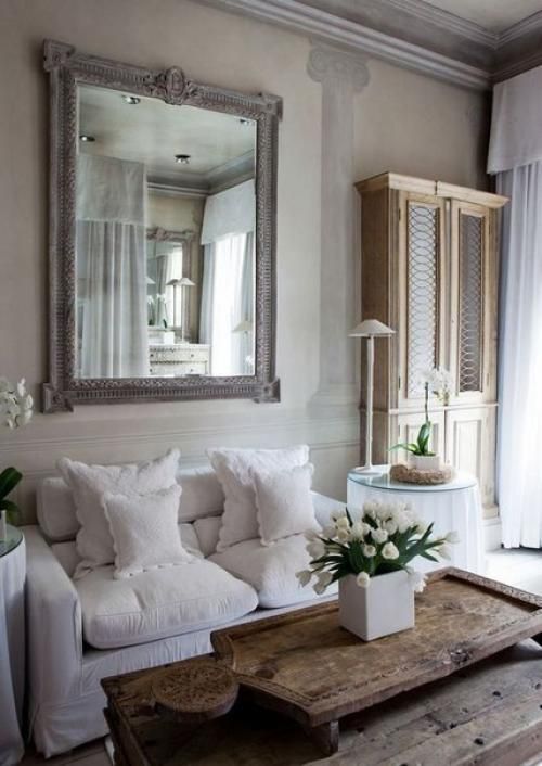 a neutral shabby chic living room with a white sofa, a large mirror, a rough wooden table and a large vintage cabinet