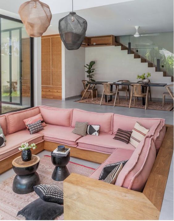 A pink conversation pit with a built in bench with pink upholstery, printed pillows, black coffee tables and a printed rug, pendant lamps
