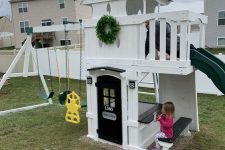 a pretty kids’ playhouse of white planks, with a slide, a black door, a built-in dining space, swings and a greenery wreath