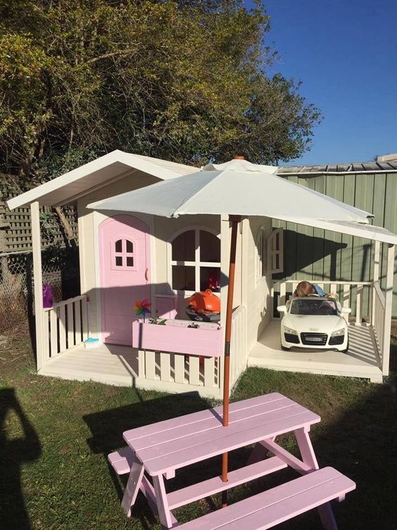 a pretty pink and neutral kids' playhouse with a pink door, potted plants, a little dining set with an umbrella and a car